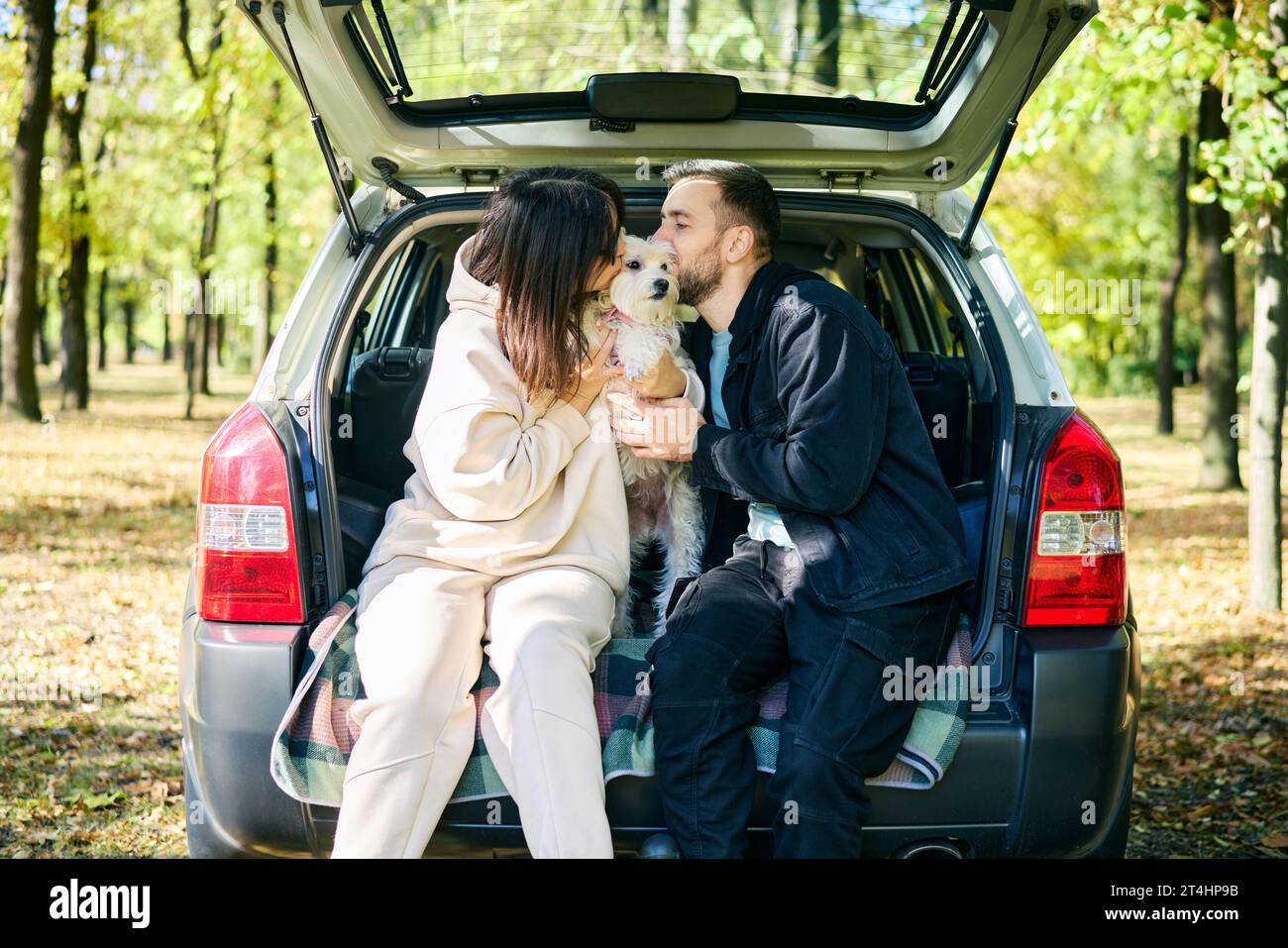 Couple affectionately kissing their lovely dog sitting in car trunk relaxing in the forest. Family, love, vacation and  travel concept Stock Photo