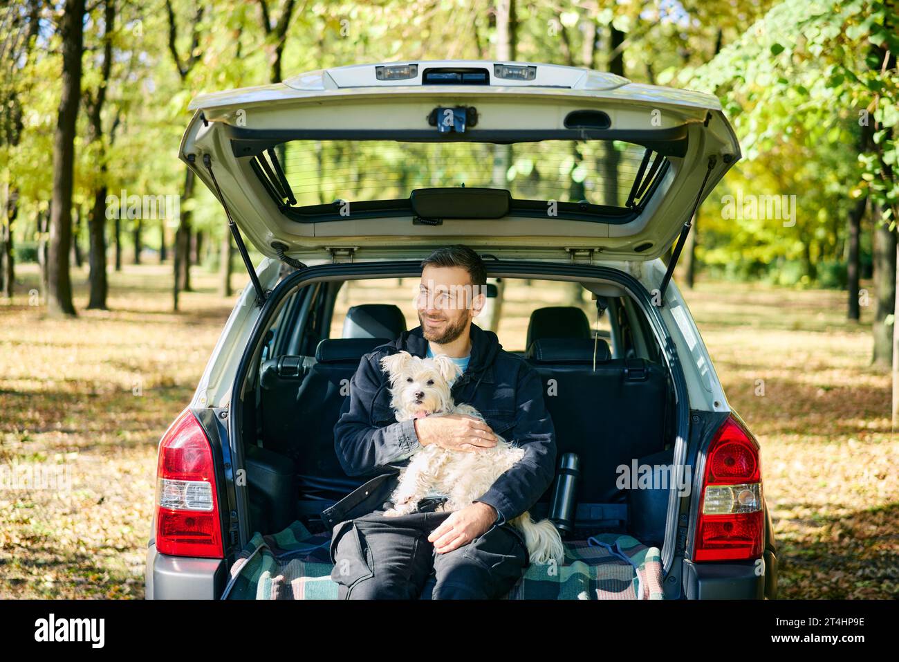Young man with a dog relaxing and enjoying nature sitting in car trunk. Vacation, holidays, travel,  adventure Stock Photo