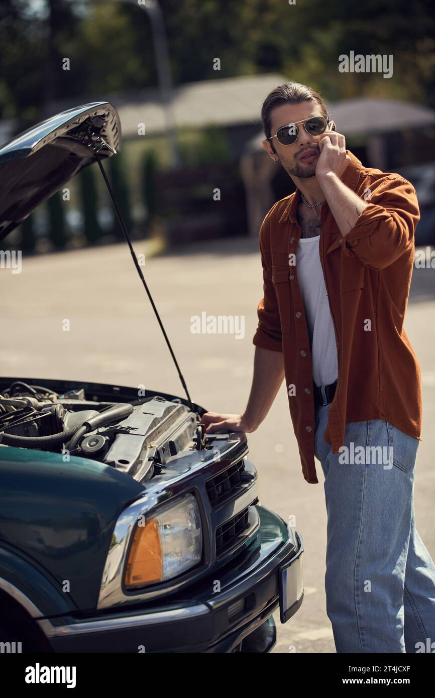 vertical shot of male driver talking by phone with his insurer near opened engine hood of broken car Stock Photo