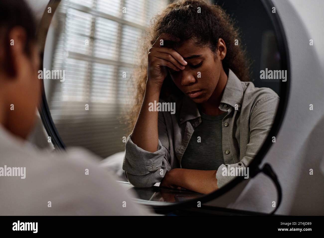 Offended teenage girl touching her forehead while sitting in front of mirror and thinking about cyberbullying messages from someone anonymous Stock Photo
