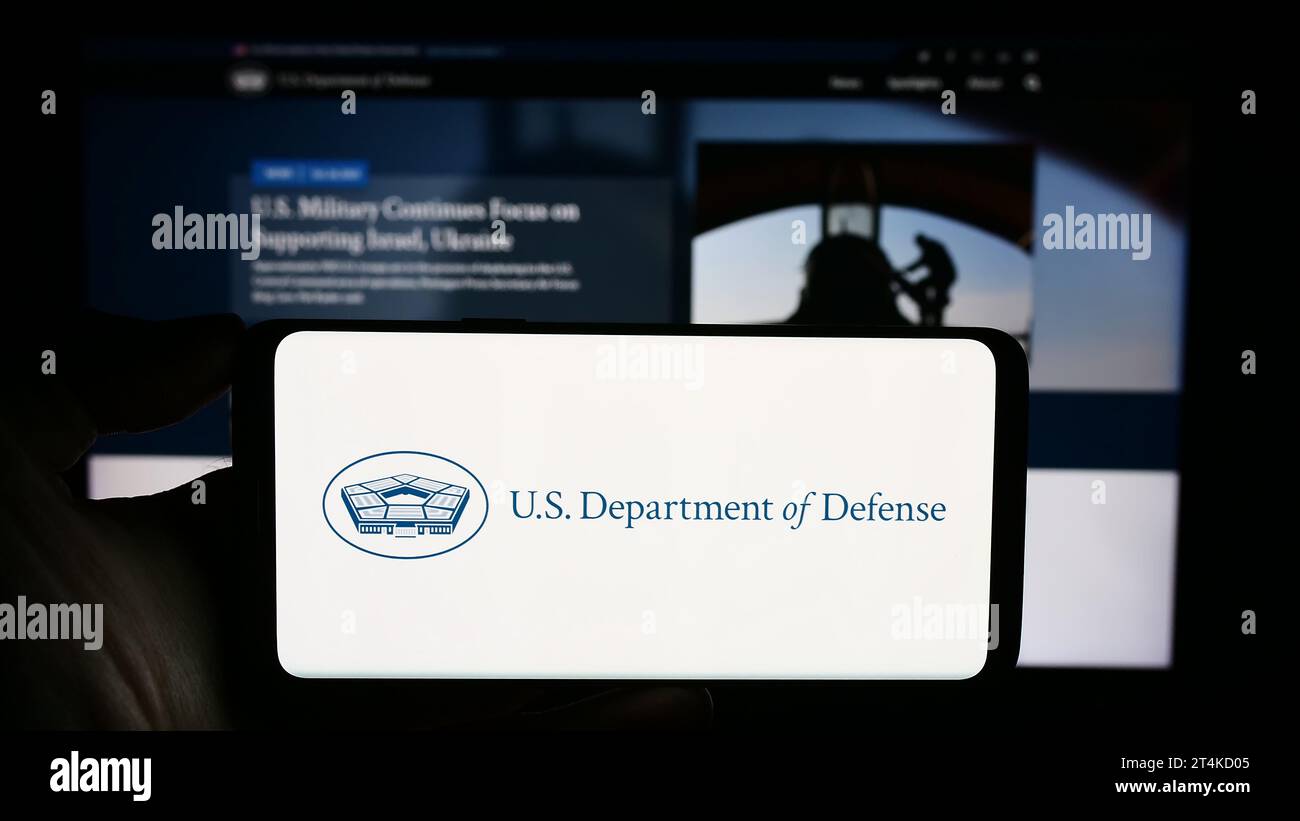 Person holding cellphone with logo of United States Department of Defense (DoD) in front of webpage. Focus on phone display. Stock Photo