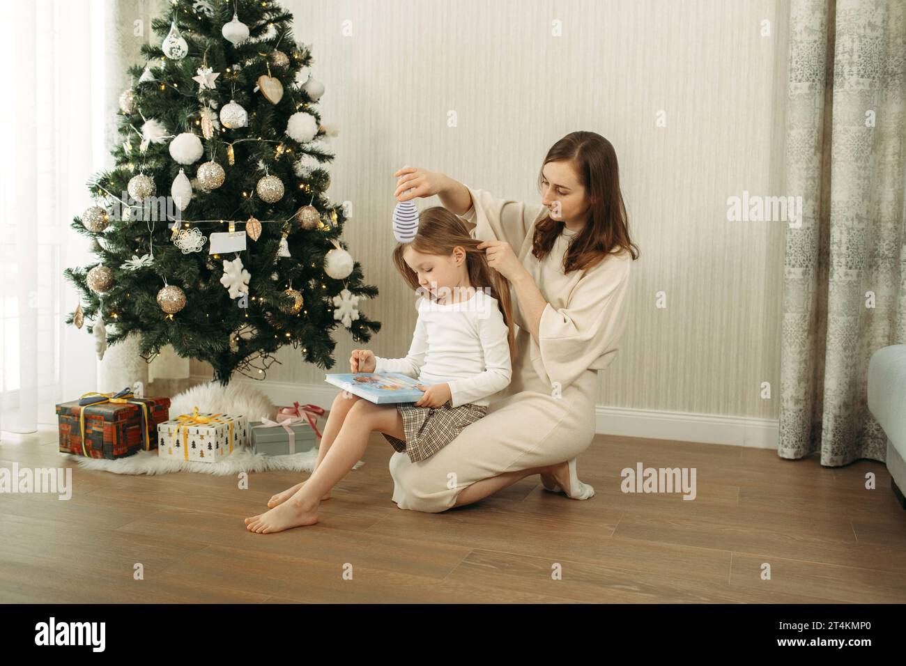 Mom combs her daughter's hair while sitting on the floor near the Christmas tree. Tender relationship between mother and daughter Stock Photo