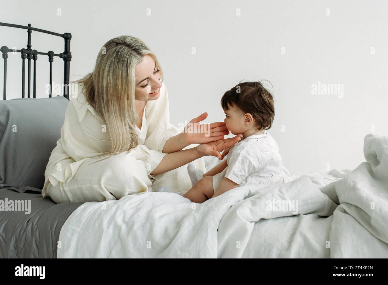 Portrait of a little girl and mother who are sitting on the bed in the morning after sleep. A happy mother holds out her hands to her daughter and smi Stock Photo