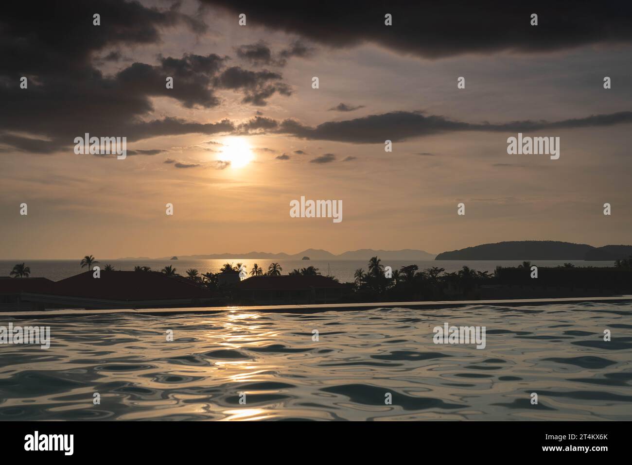 Dramatic tropical evening Sun in Thailand - view over roof top swimming pool in luxury hotel. Dark clouds over palms, sea and mountains at horizon Stock Photo