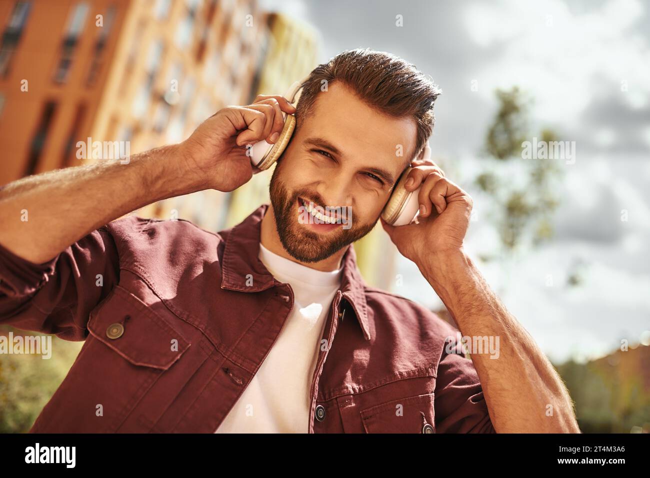 Can't live without music. Handsome and happy young man with stubble in headphones listening to the music and smiling while standing on the street Stock Photo