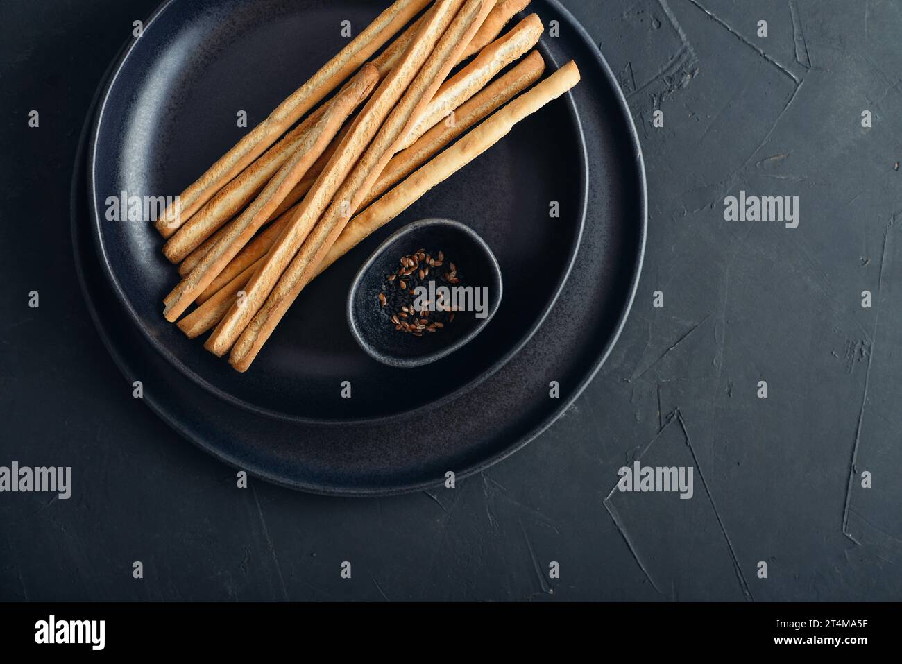 Fresh baked Italian grissini breadsticks on plate with sesame and flax seeds on black background, top view Stock Photo