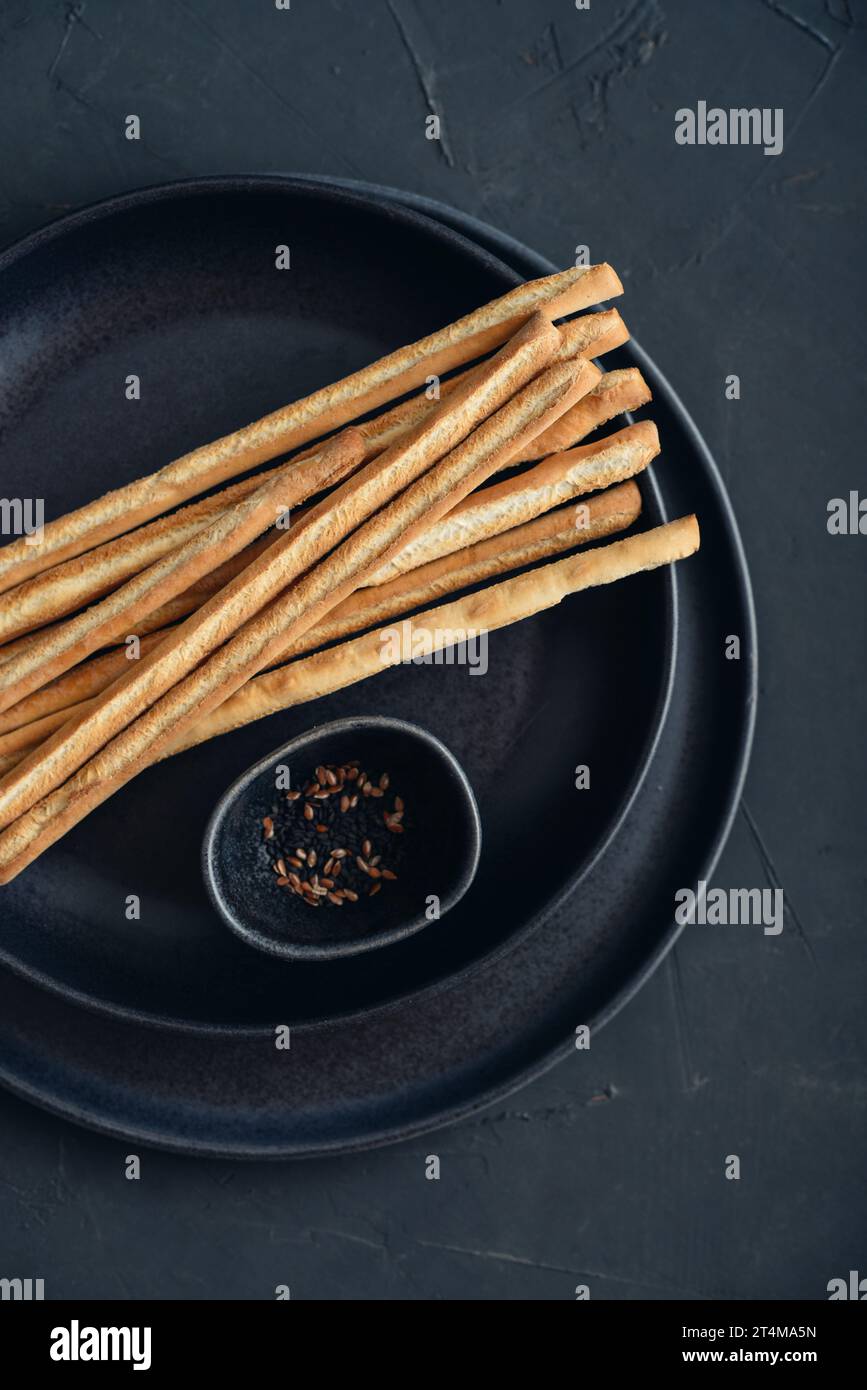 Fresh baked Italian grissini breadsticks on plate with sesame and flax seeds on black background, top view Stock Photo