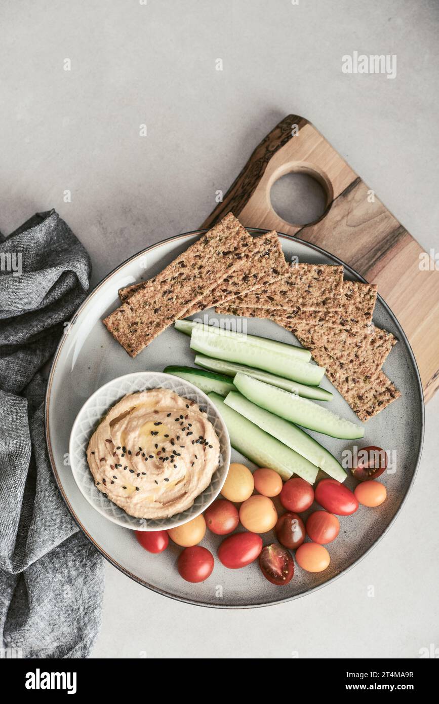 Bowl with hummus, cucumber, cherry tomatoes and crispbread on a platter on a light background, top view Stock Photo