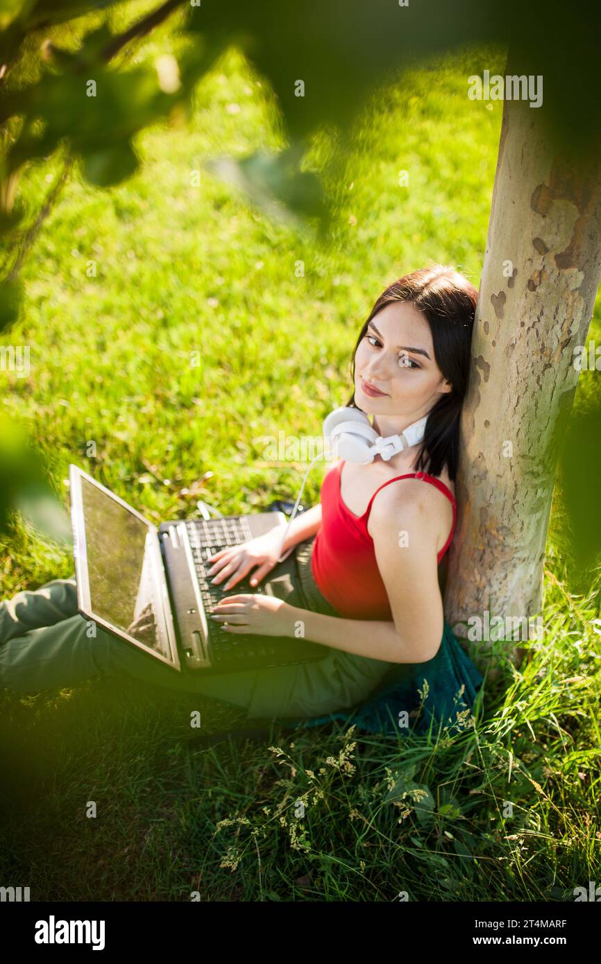 girl with dark brown hair wearing white headphones using laptop in a park under a tree Stock Photo