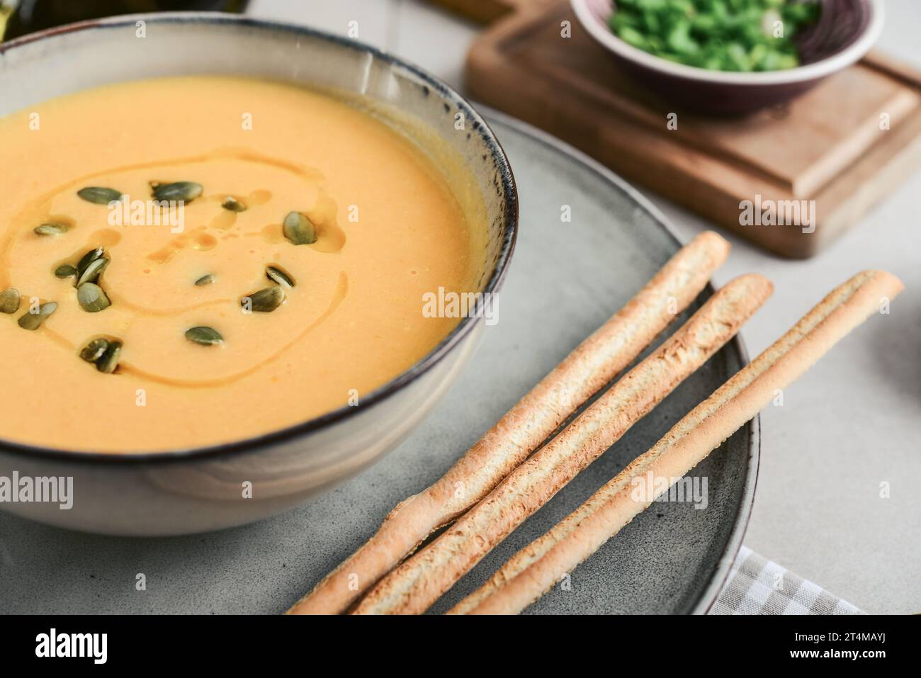Pumpkin soup in a bowl with pumpkin seeds, green onions and breadsticks on a light background, closeup Stock Photo