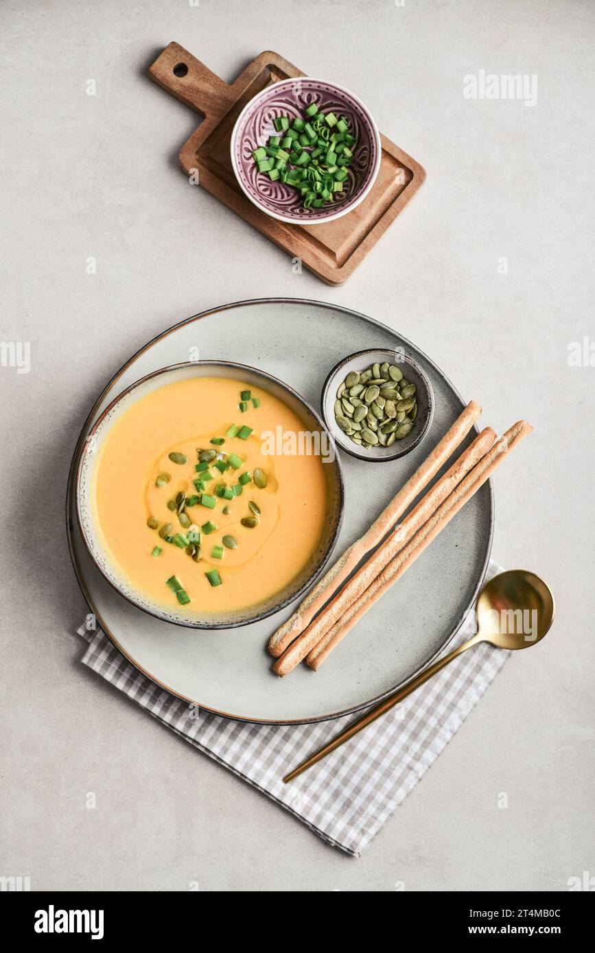 Pumpkin soup in a bowl with pumpkin seeds, green onions and breadsticks on a light background, top view Stock Photo