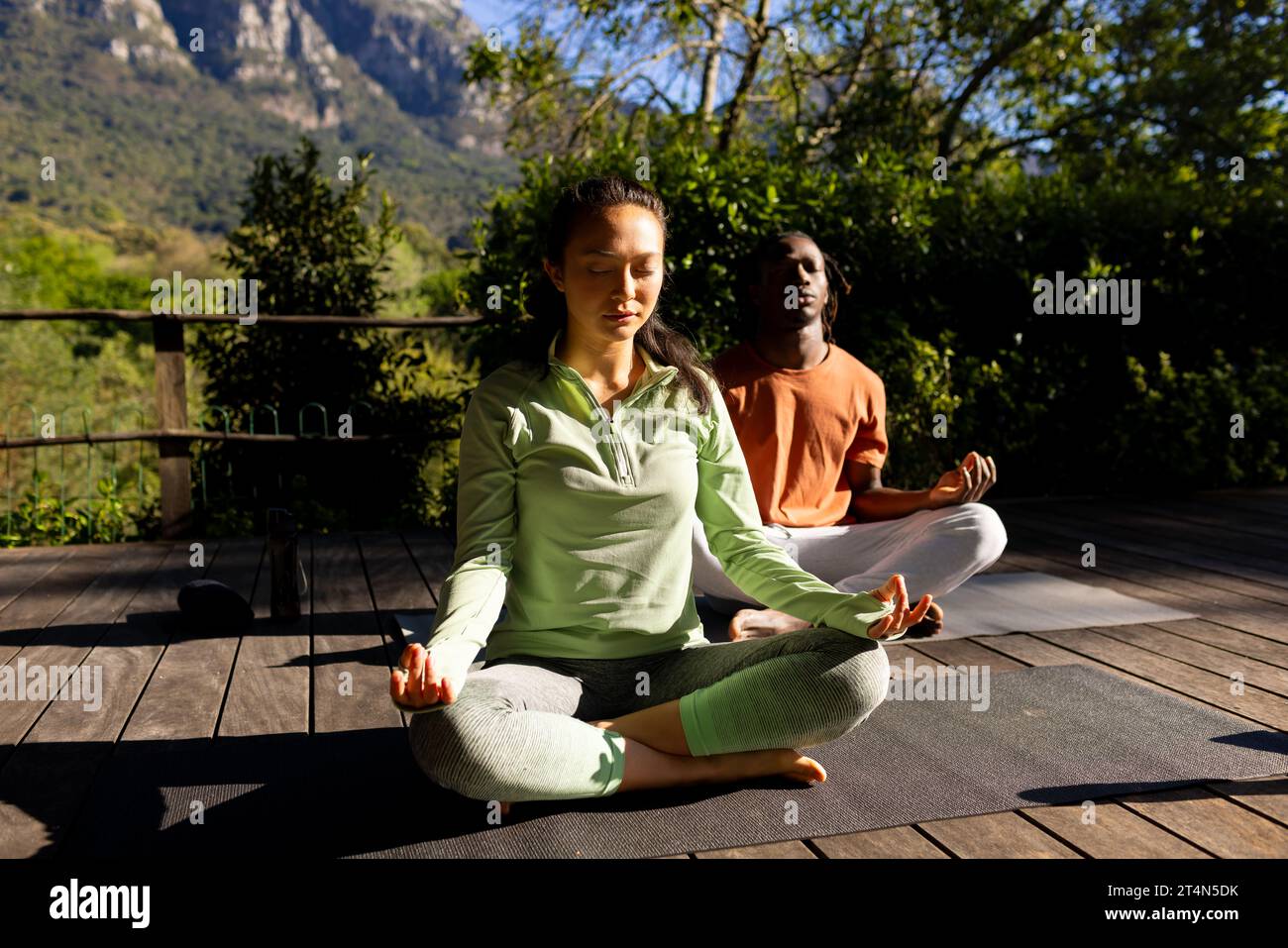 Happy diverse couple practicing yoga meditation sitting on deck in sunny garden Stock Photo