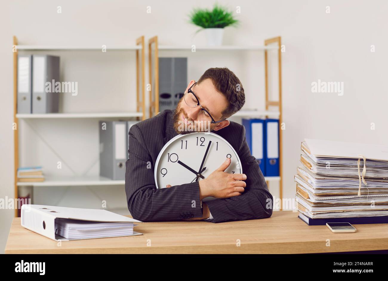 Tired, overworked office worker sitting at desk with paperwork, holding clock and sleeping Stock Photo