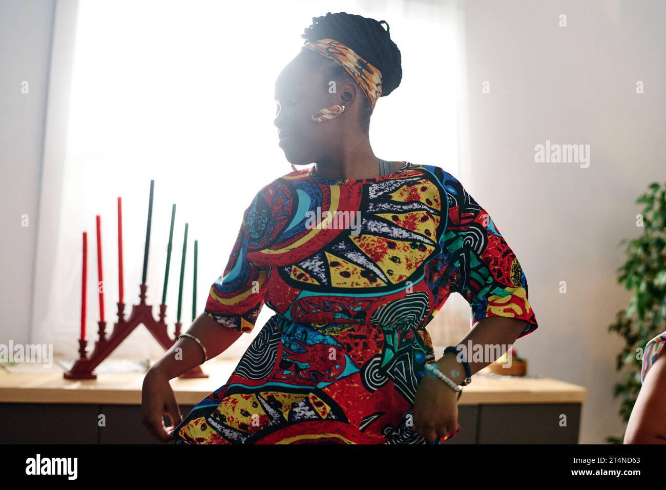 Young African American woman in ethnic attire and accessories performing national dance by table with seven candles symbolizing main principles Stock Photo