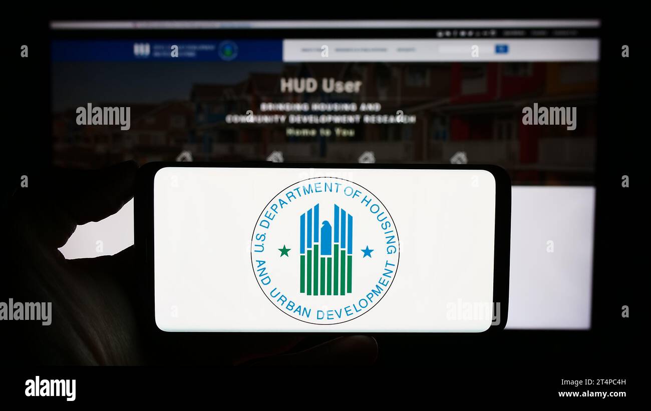 Person holding mobile phone with logo of US Department of Housing and Urban Development (HUD) in front of web page. Focus on phone display. Stock Photo