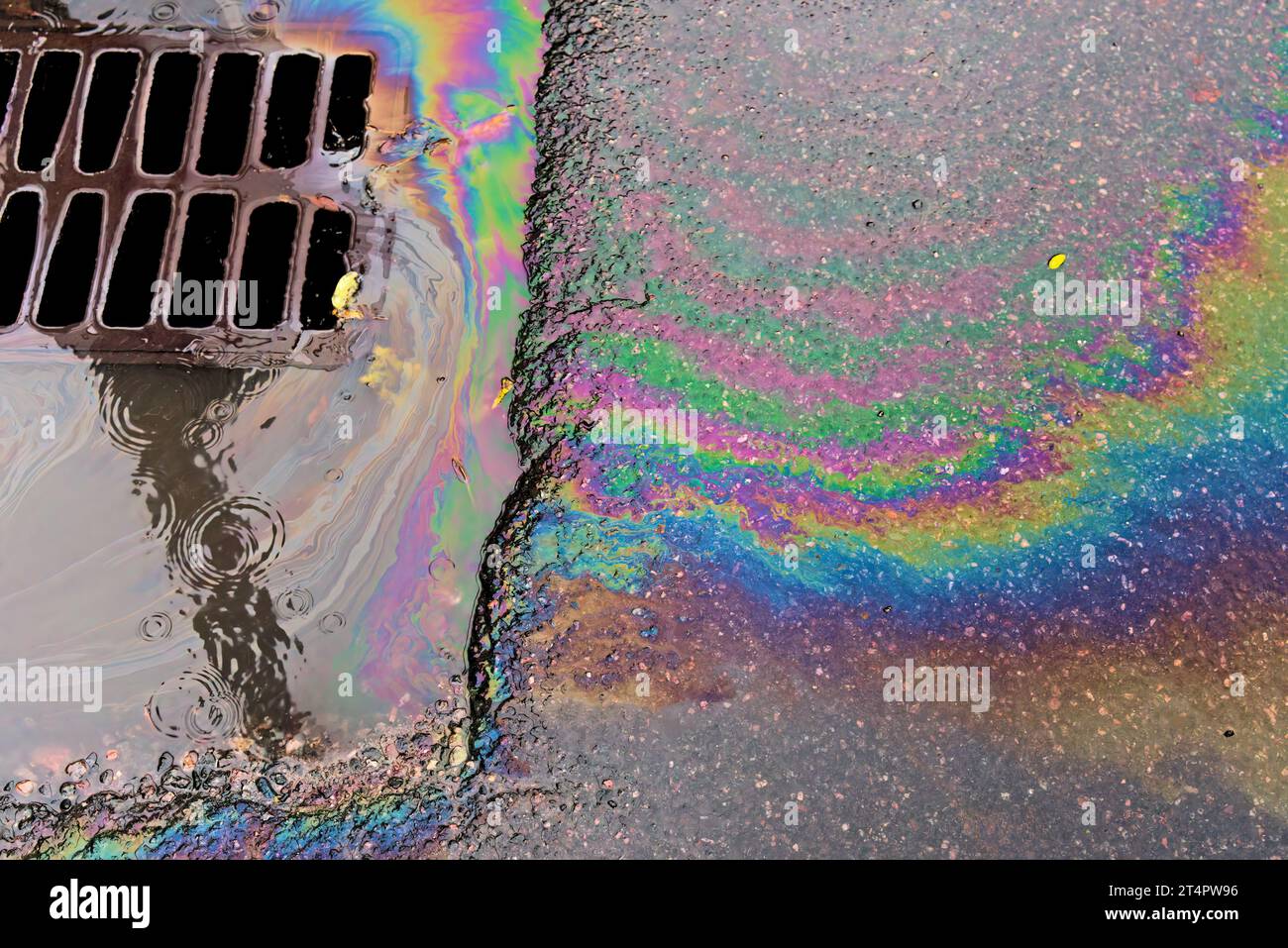An oil slick against the backdrop of an asphalt road flows into a storm drain through a grate. Environmental problems of water pollution. Stock Photo