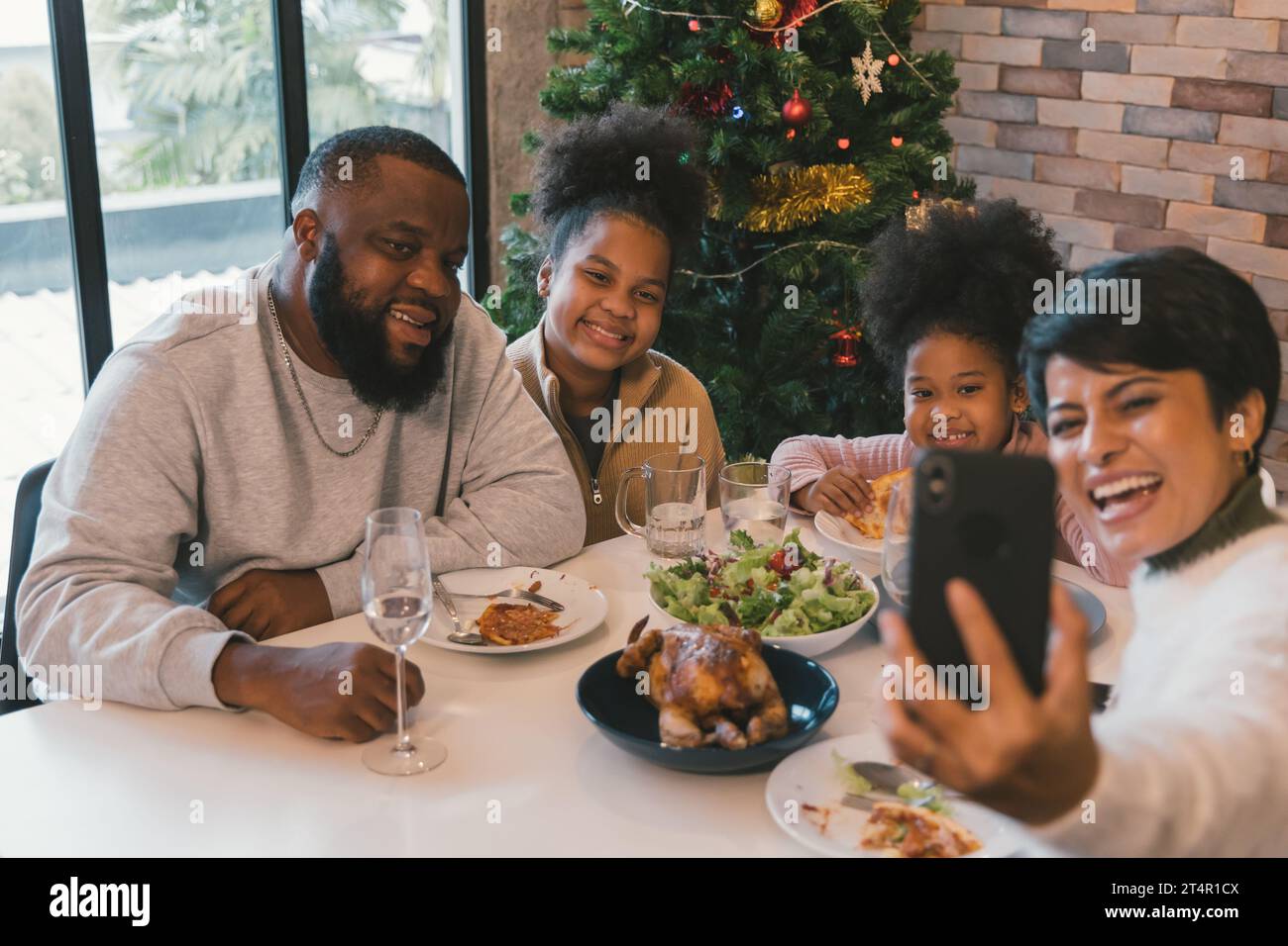 Cheerful african american family enjoying christmas and new years lunch while woman taking selfie on smartphone with members of family at home against decorated xmas tree Stock Photo