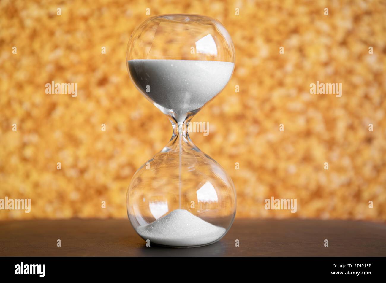 Hourglass with white sand dripping against gold color glitter background indoors. Stock Photo