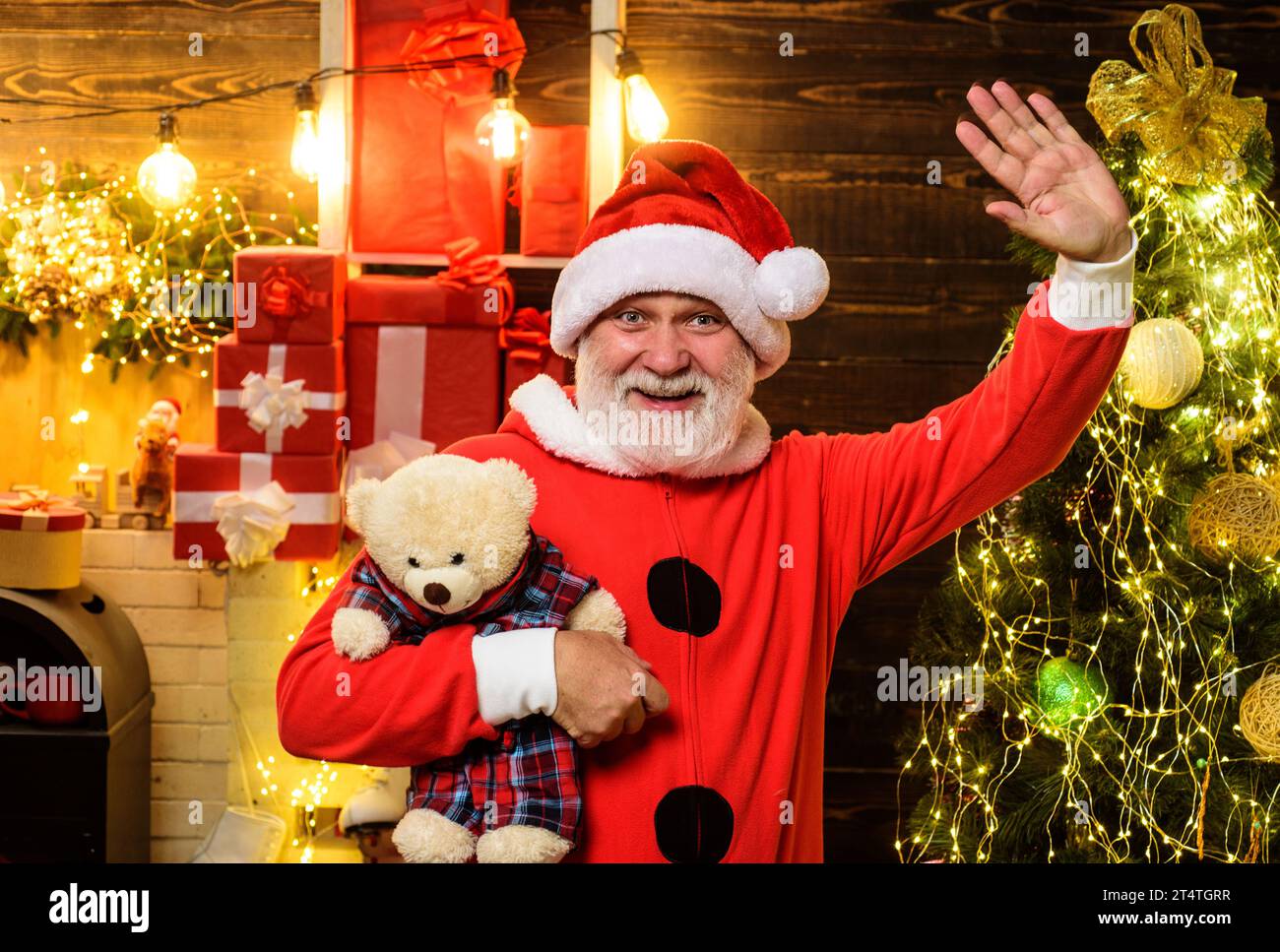 Christmas or New Year holidays. Bearded man in Santa Claus costume with teddy bear toy waving hand. Smiling Santa Claus with teddy bear in room Stock Photo
