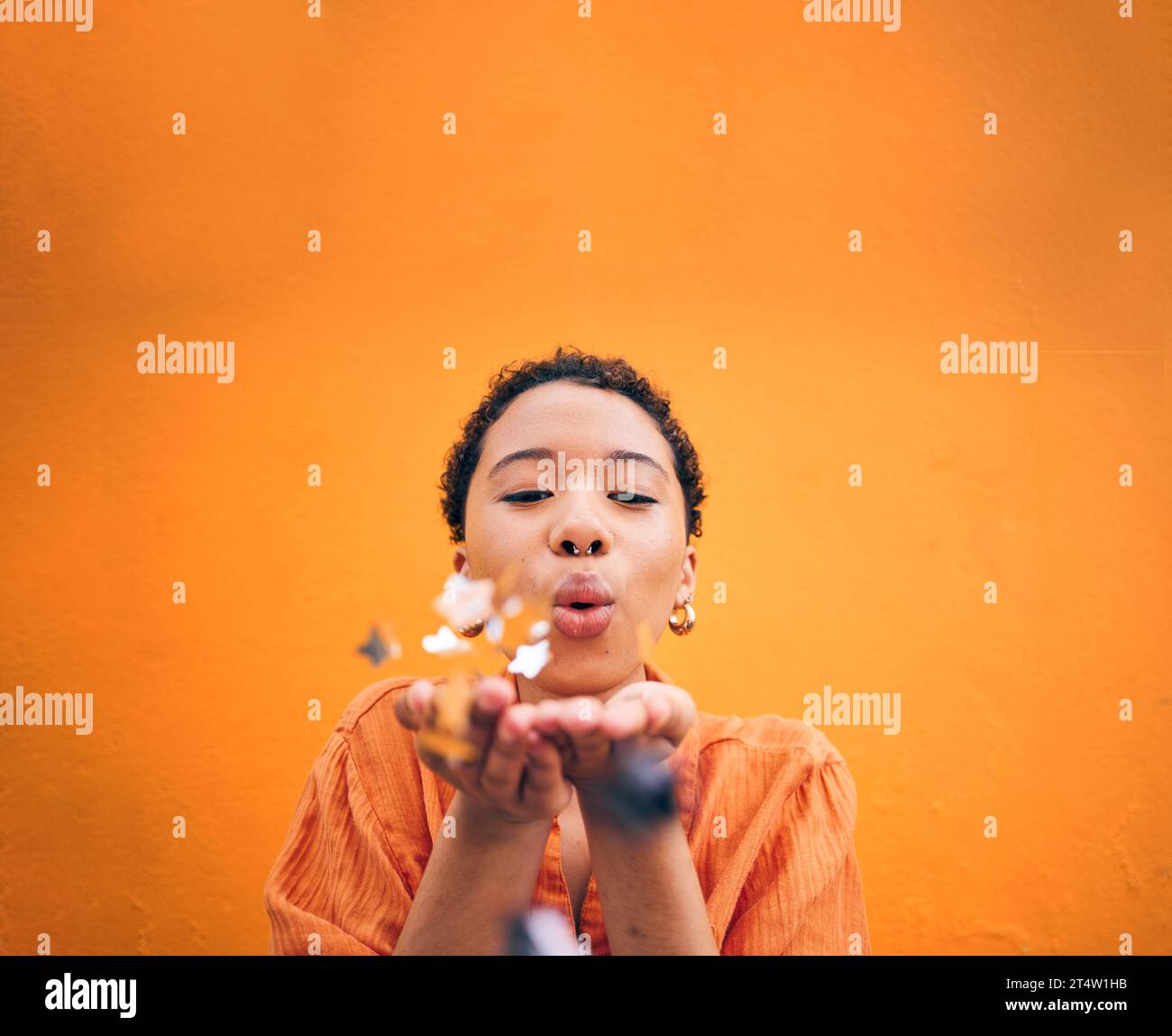 Blowing, petals and face of woman on orange background for natural beauty, skincare and romance. Flowers, happy and female person with floral confetti Stock Photo