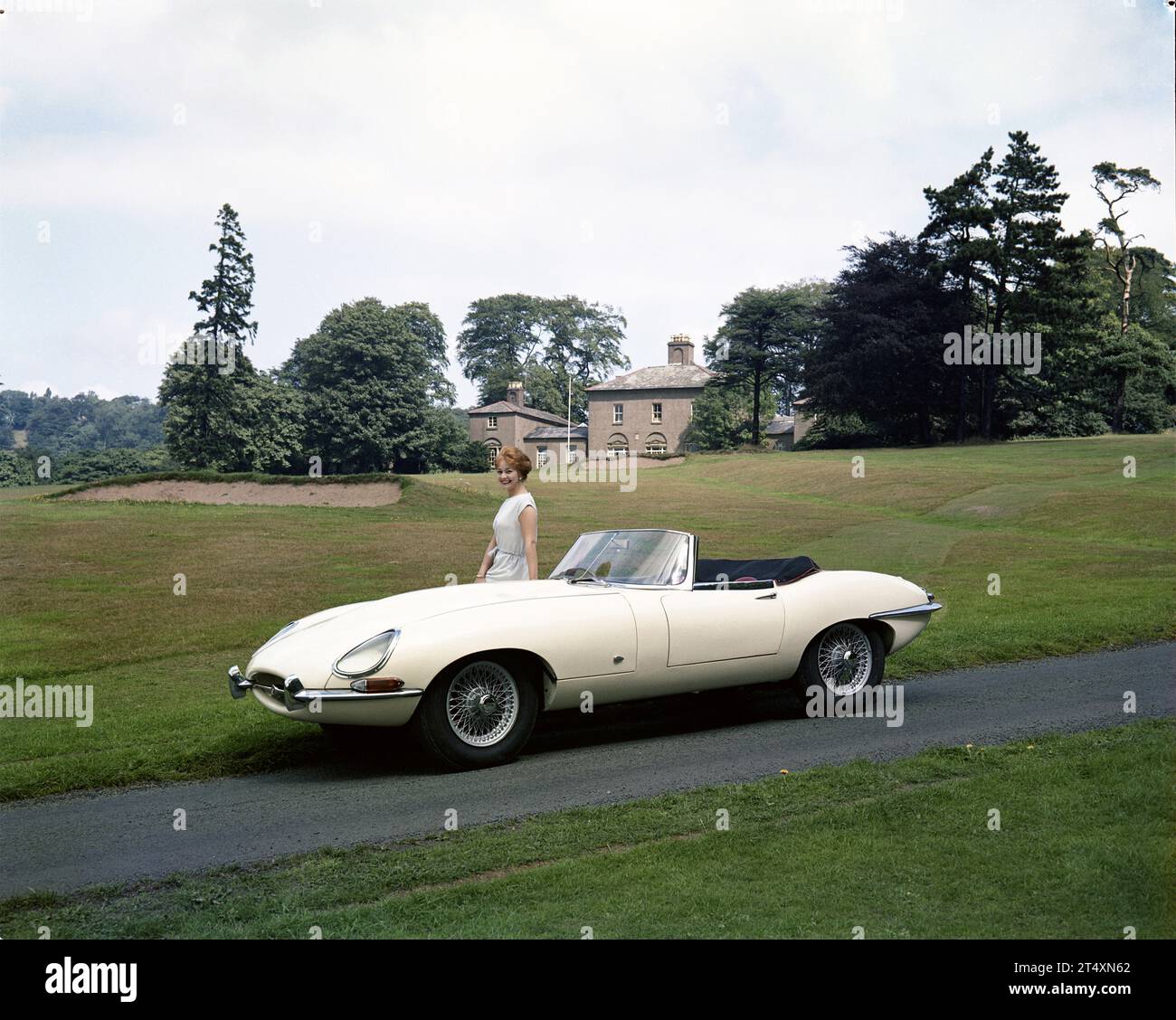 Early publicity photo of Jaguar E-type S1, Open Two-Seater, 3.8L, with model in white dress at Halesowen Golf Club with clubhouse in the background. Stock Photo