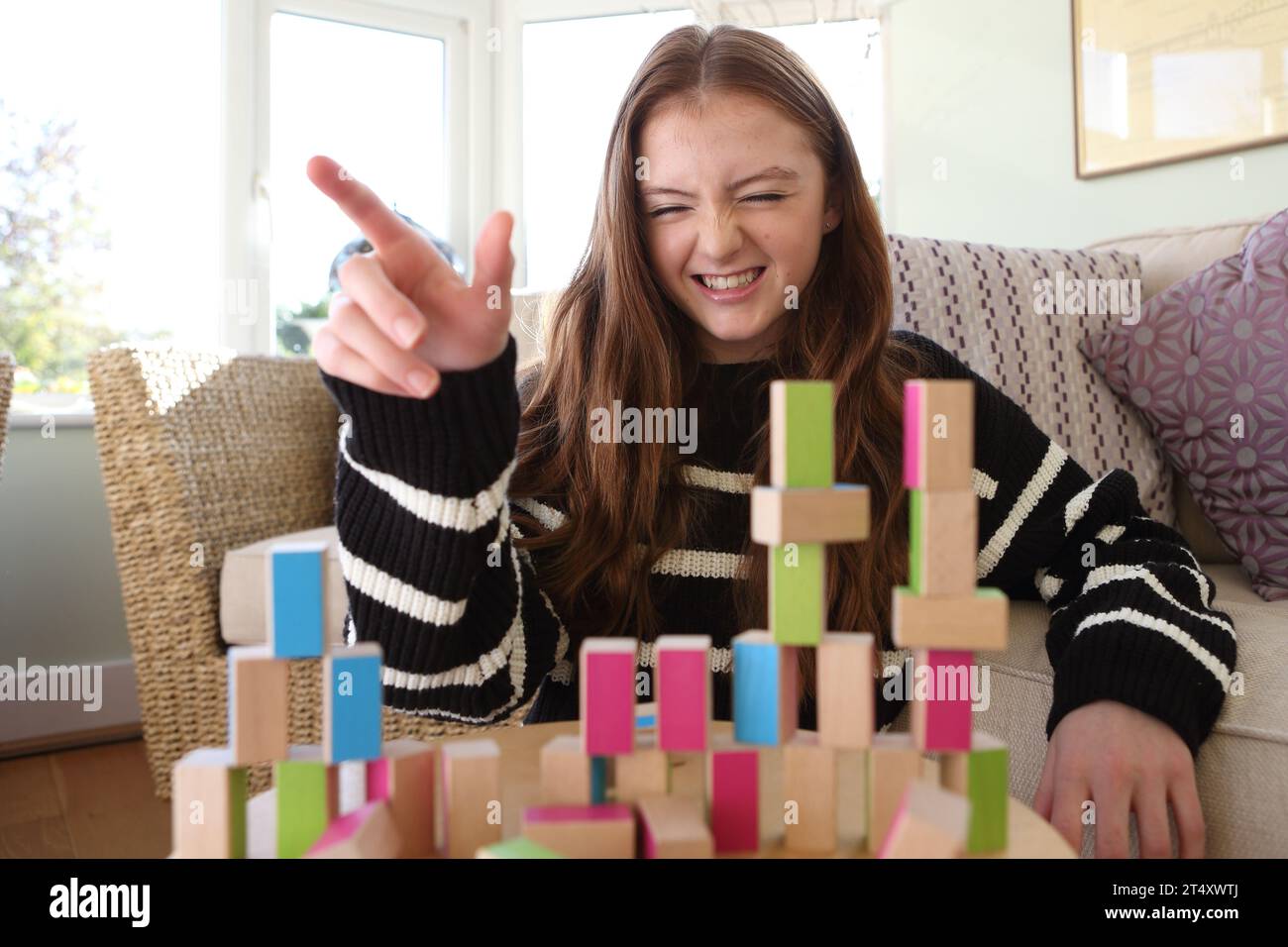 Teenage girl smiling and laughing destroying a wall and towers of wooden blocks, knocking down the pattern with her hand Stock Photo