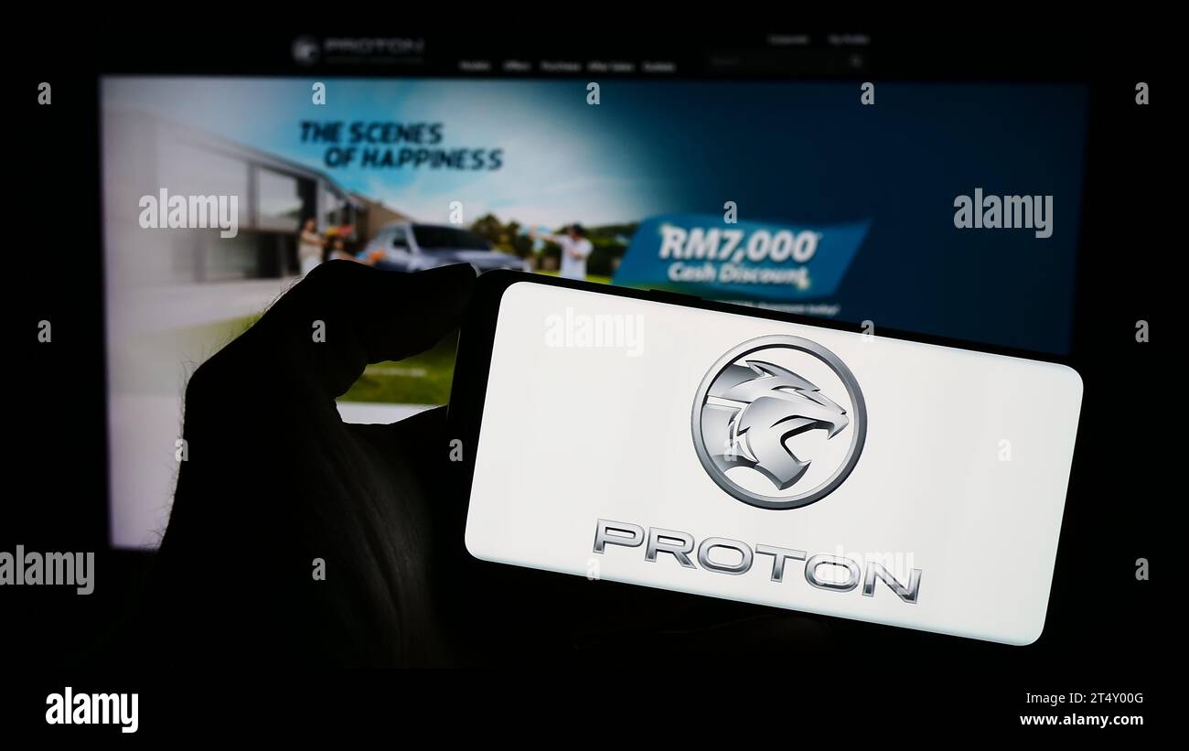 Person holding cellphone with logo of Malaysian automotive company Proton Holdings Berhad (PHB) in front of webpage. Focus on phone display. Stock Photo