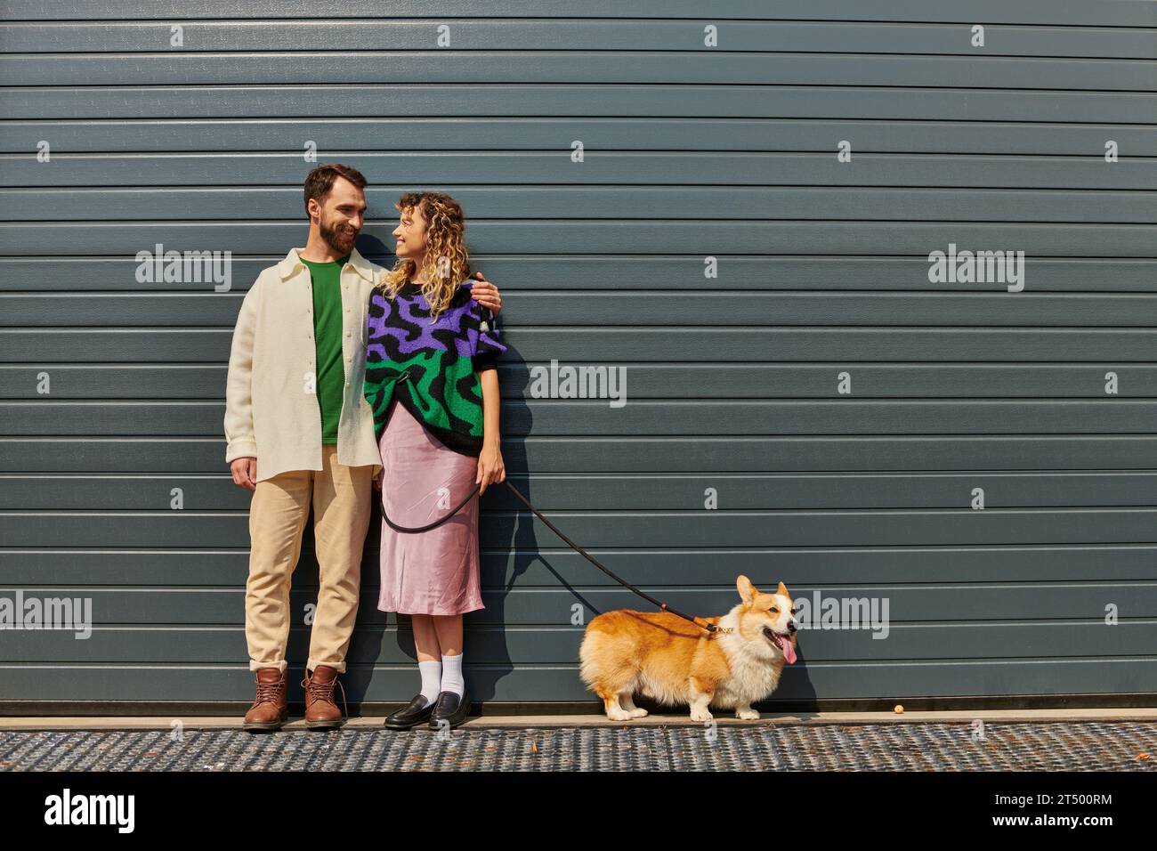 cheerful and stylish couple walking with corgi dog near grey garage door, animal companions Stock Photo