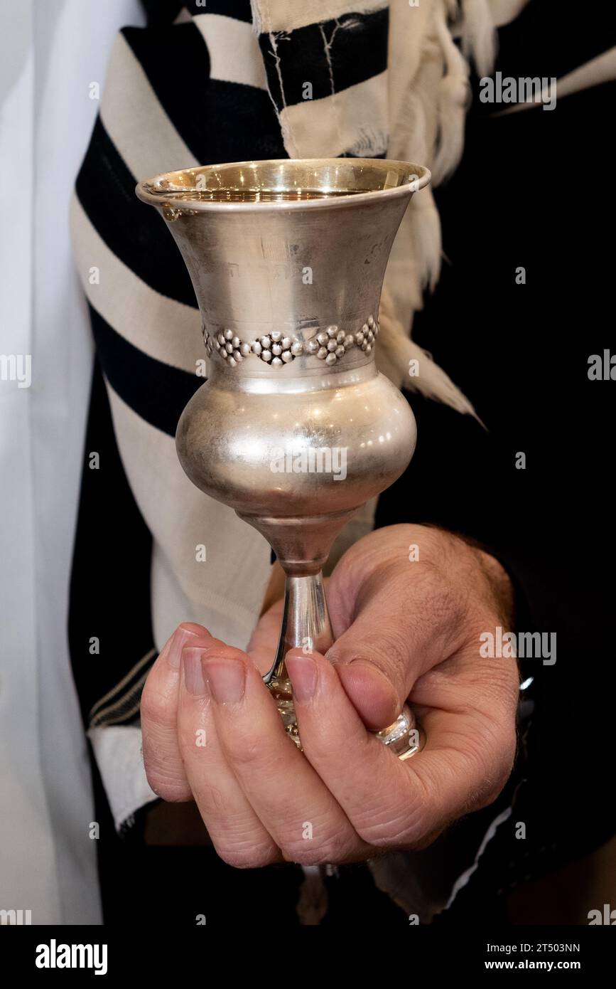 A man holds a silver, decorative cup of wine in one hand and recites a blessing for the bride and groom under the chuppah or wedding canopy at a Jewis Stock Photo