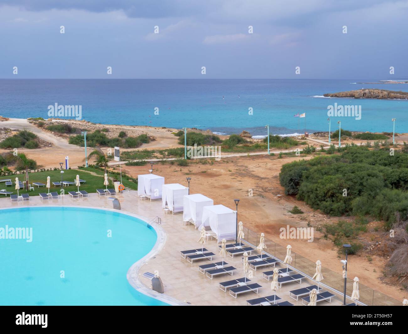 Top view over empty swimming pool with sun beds and umbrellas, Mediterranean sea with small island near Nissi beach and path promenade along the shore Stock Photo