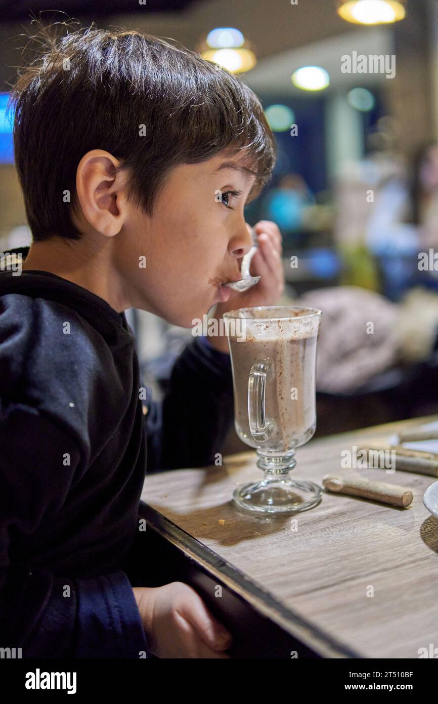 Latino brunette little boy in profile in a bar drinking hot chocolate milk with a spoon. Background with blurred lights Stock Photo