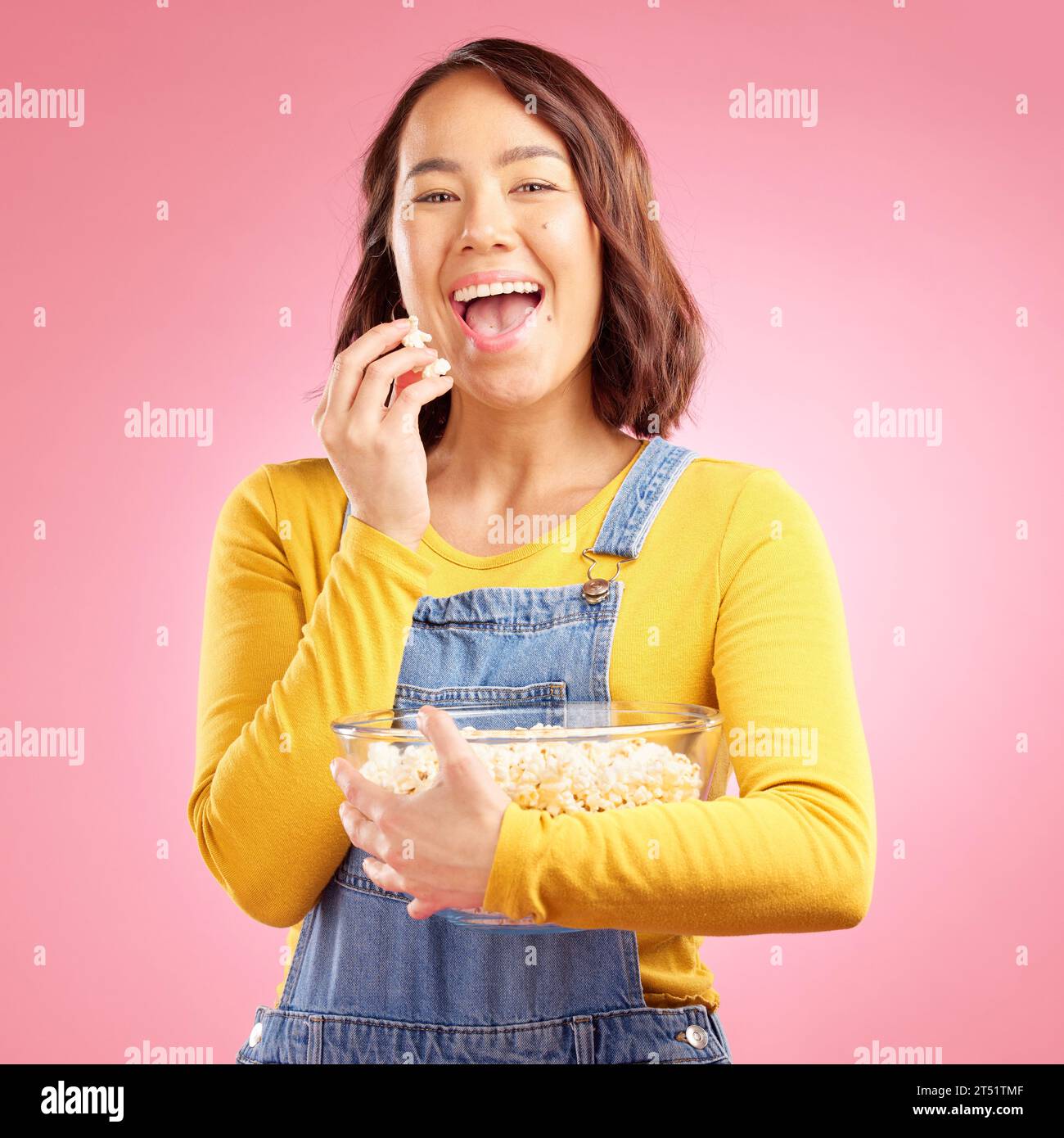 Portrait, asian woman and eating popcorn in studio while watching movie, film or cinema show on pink background. Happy female model, bowl or snack to Stock Photo