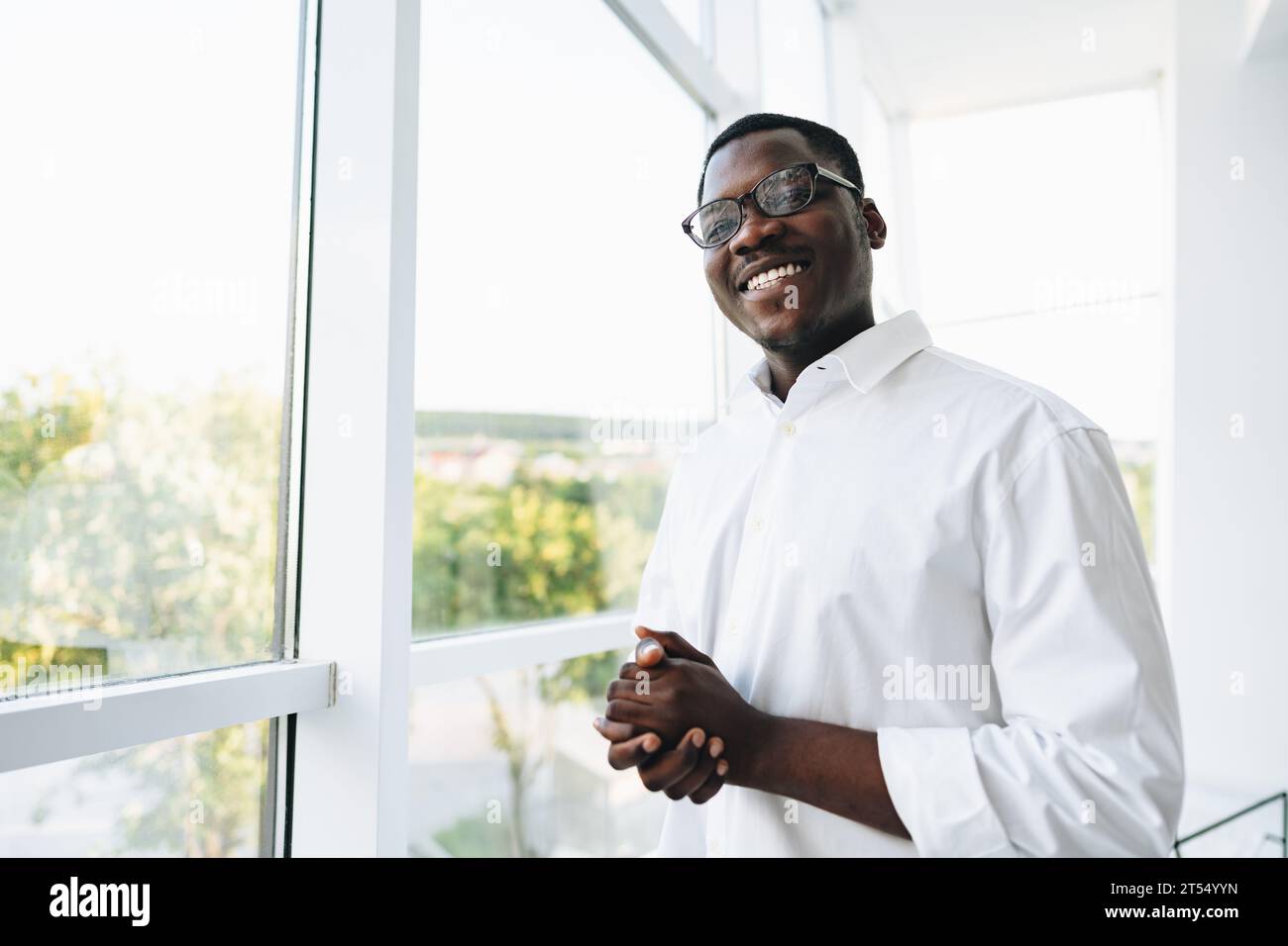 Young african man in white shirt and glasses posing near large window ...