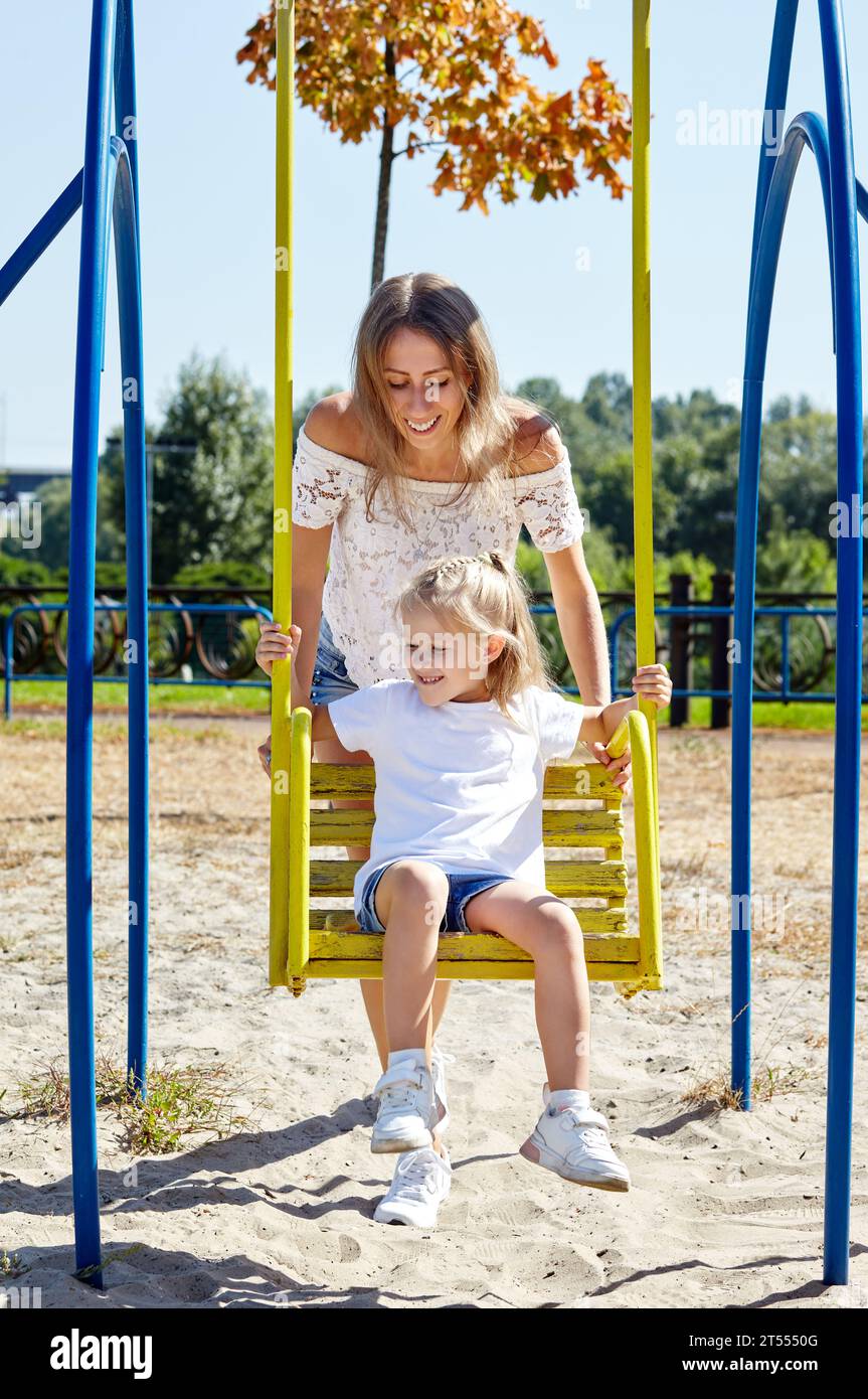 Mother with daughter on swing on a playground in the summer city park. Childhood, leisure and people concept - happy family rest and have a good time Stock Photo