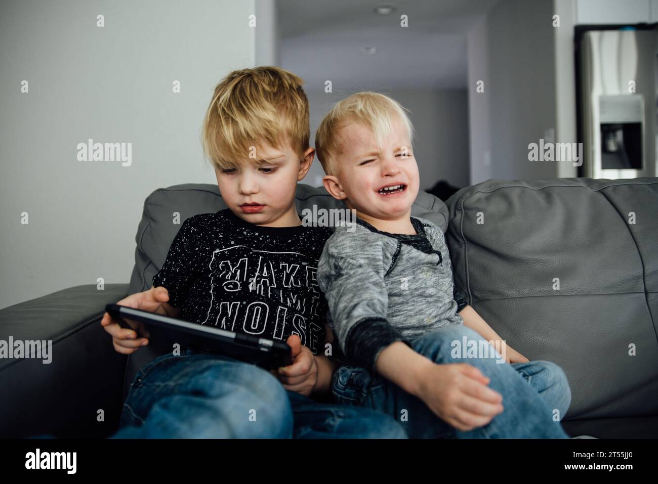 Two boys sit on a couch while one holds an ipad and the other th Stock Photo