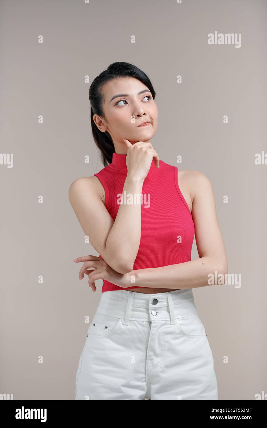 Asian thoughtful woman keeps hand on chin looks pensively against beige ...