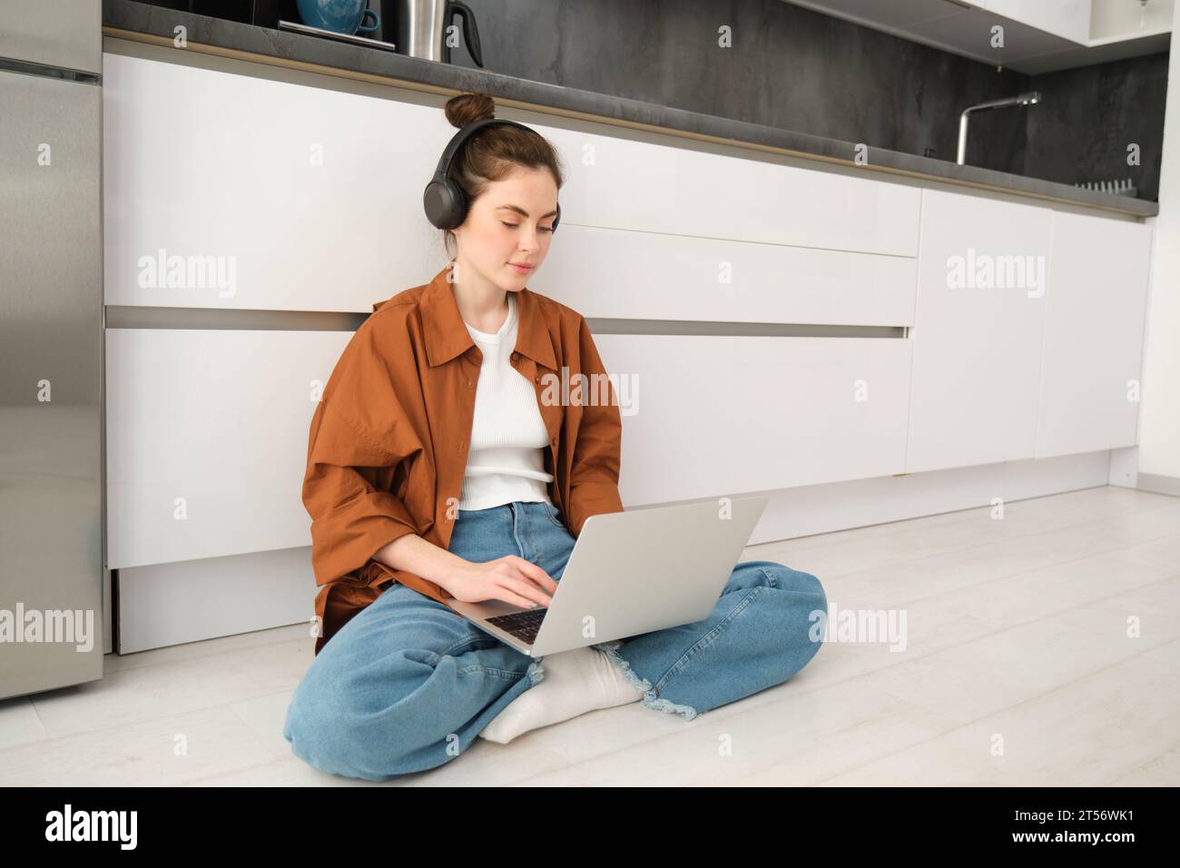 Young woman freelancer, working from home, student sitting with laptop on kitchen floor, wearing headphones, typing on computer. Stock Photo