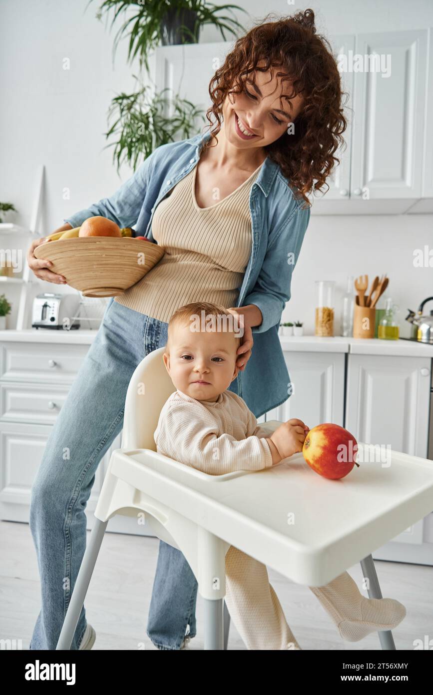 joyful mother with fresh fruits stroking head of baby daughter sitting in baby chair near ripe apple Stock Photo