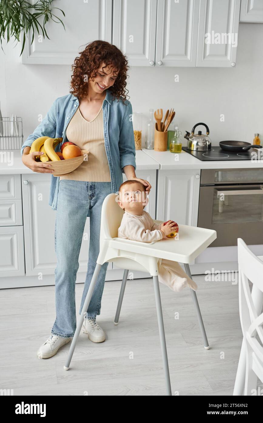 smiling woman with fresh fruits stroking head of baby girl sitting in baby chair, morning in kitchen Stock Photo