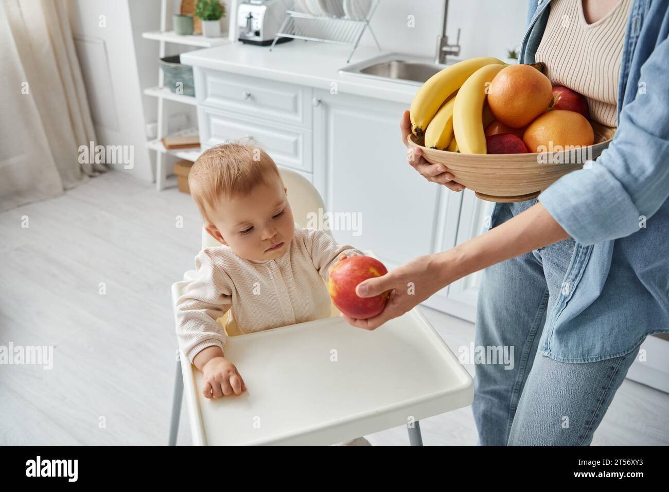 mother with bowl of fresh fruits proposing ripe apple to little daughter sitting in baby chair Stock Photo