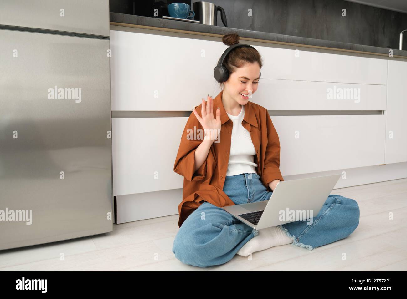 Remote workplace. Young woman works from home, sits on floor with headphones and laptop, student doing homework, studying online. Stock Photo