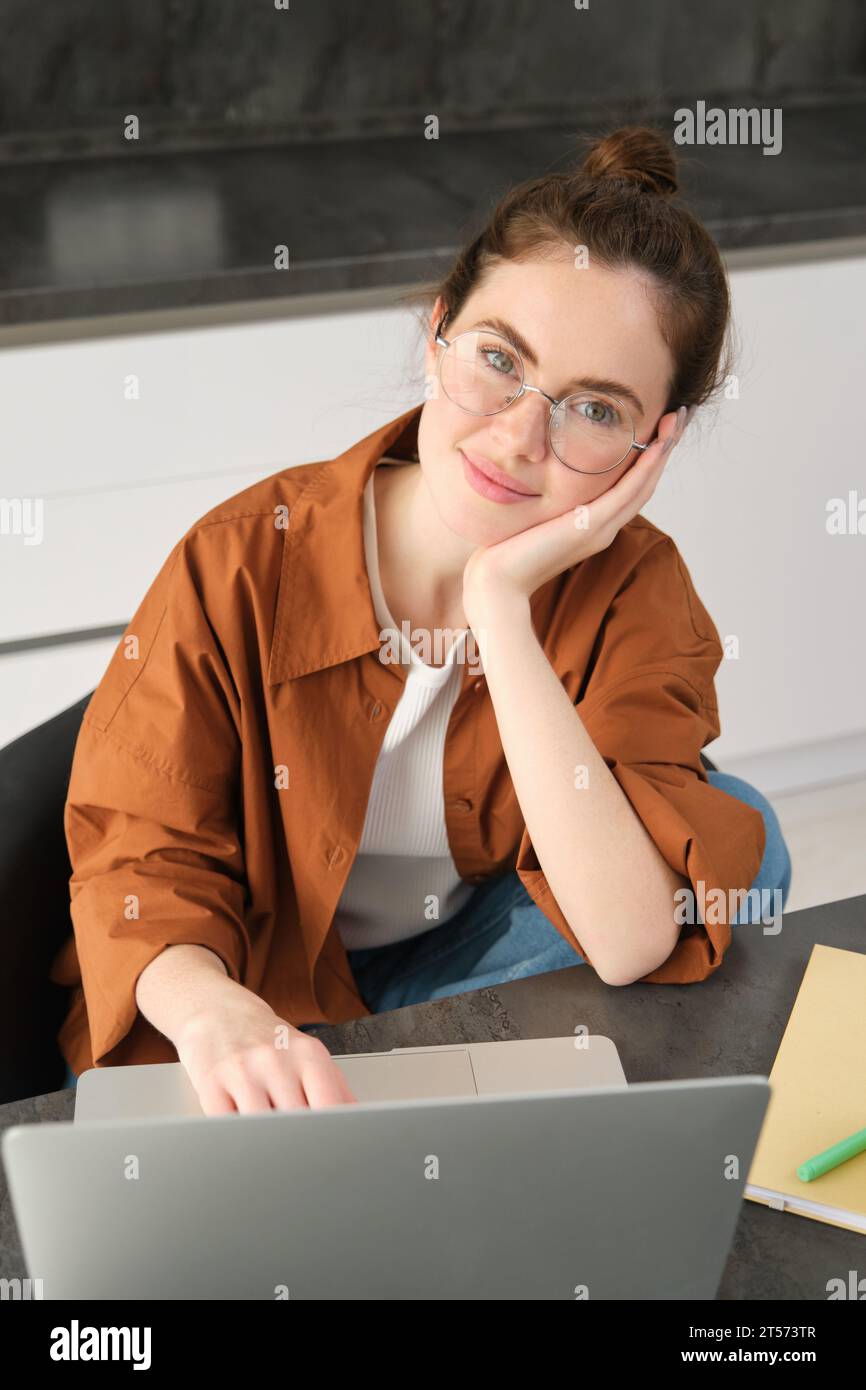 Vertical shot of beautiful woman working from home, student doing homework in kitchen, using laptop, looking at camera and smiling. Stock Photo