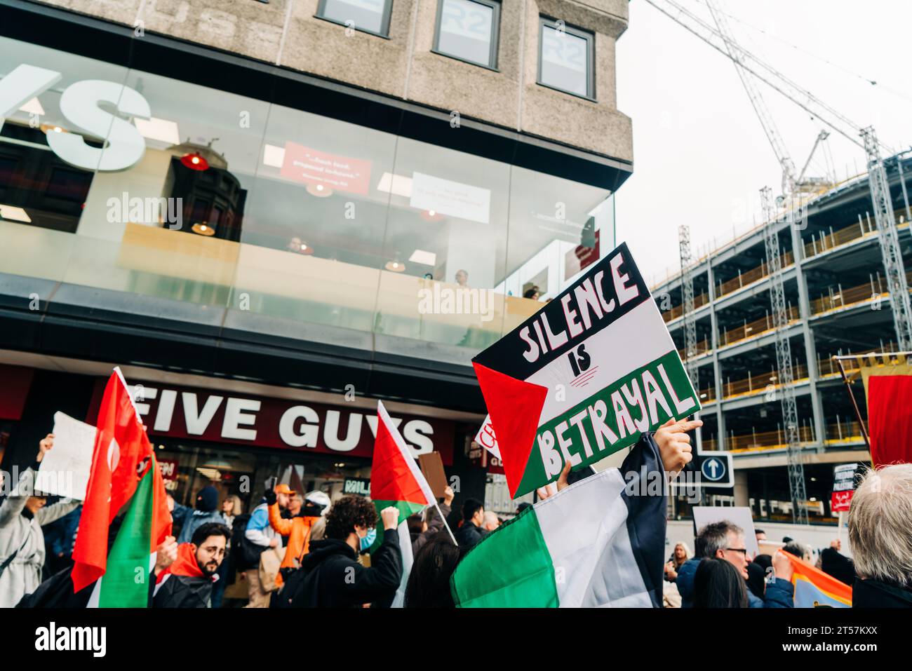 Protester holds up sign reading 'Silence is betrayal' written on Palestine flag in front of Five Guys. Newcastle Upon Tyne, England, UK - Oct 28 2023. Stock Photo