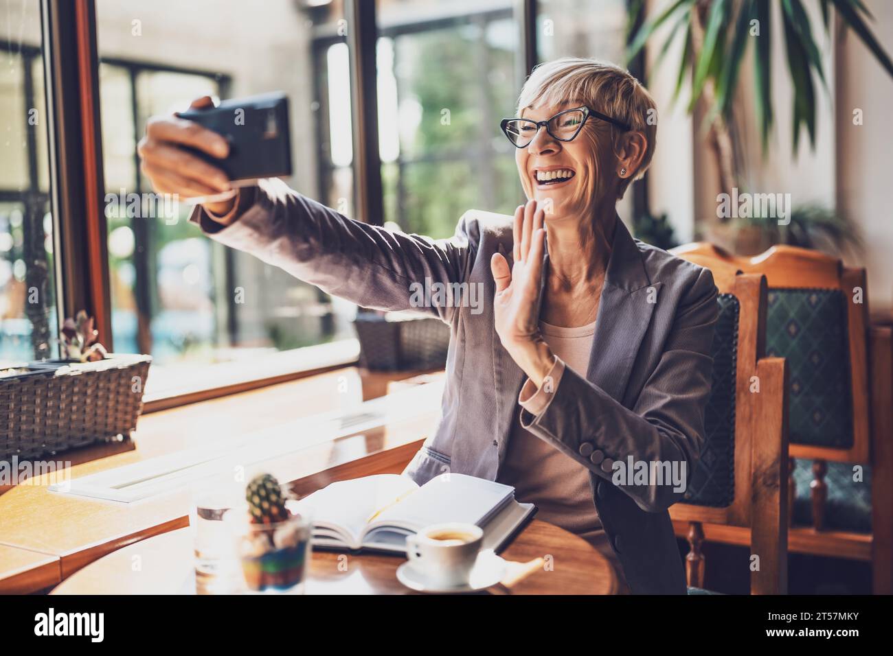 Mature woman is sitting in cafe and relaxing. She is drinking coffee and using smartphone. Stock Photo