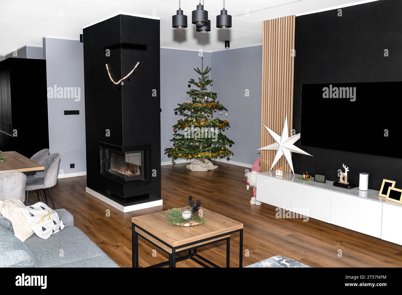 A Christmas tree made of Caucasian fir decorated with baubles stands in the hall of a modern house, with a fireplace visible in the living room. Stock Photo