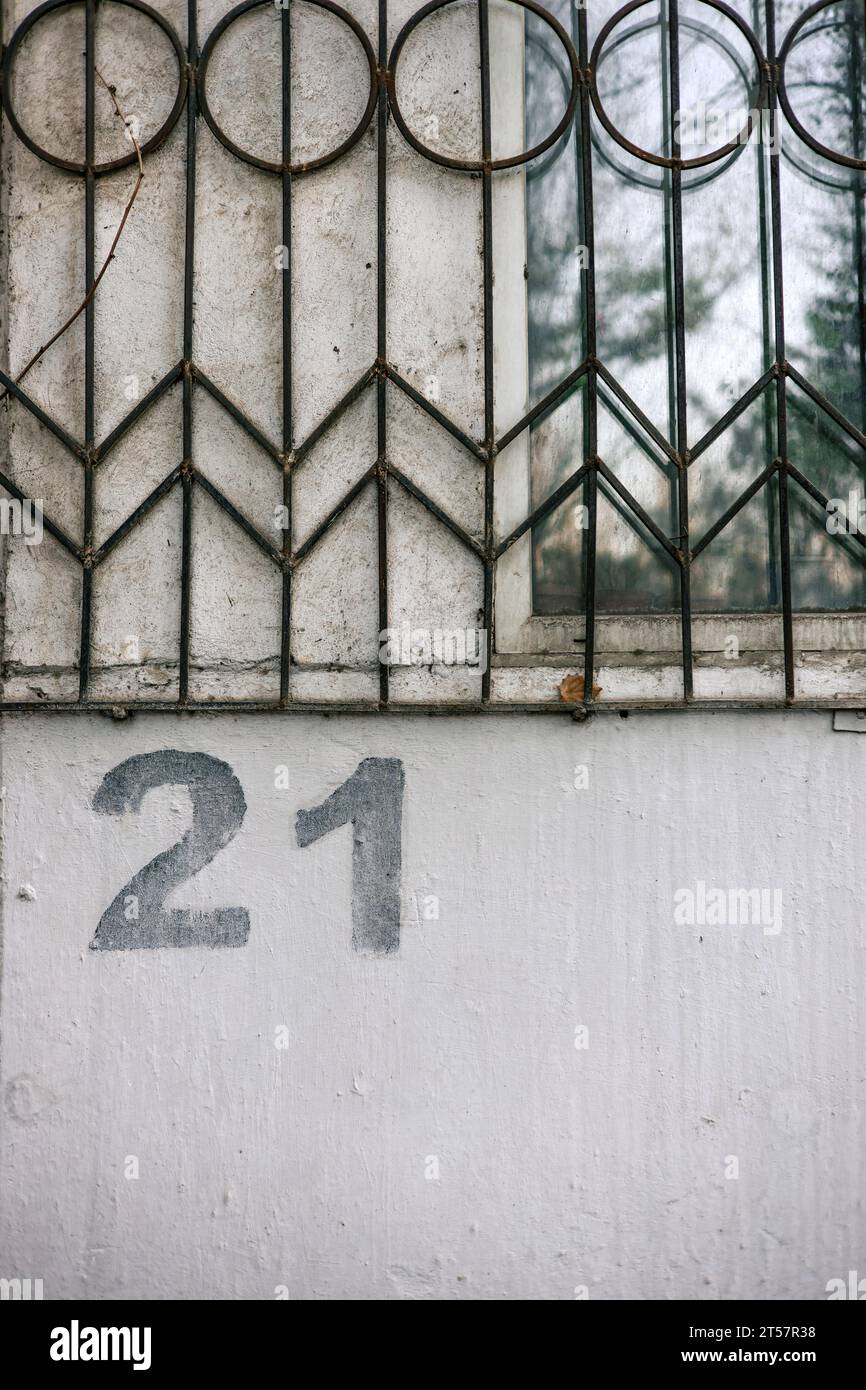 The number twenty one is painted using a stencil on a gray wall below a window with bars. House numbering 21 Stock Photo
