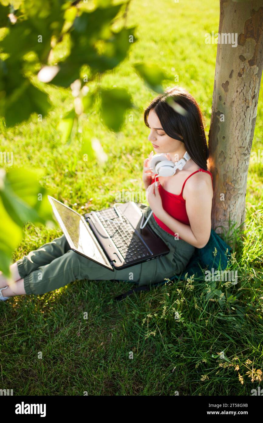girl with dark brown hair wearing white headphones using laptop in a park under a tree Stock Photo