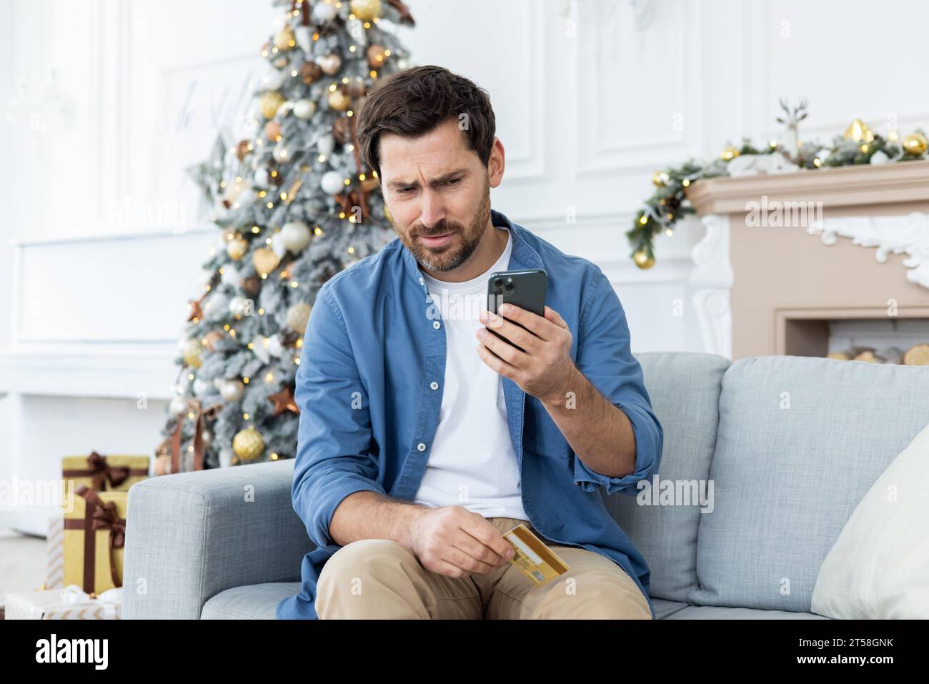 Credit score problems. A mature man is sitting at home on the sofa on Christmas holidays and holding a credit card in his hands, looking at the phone screen incredulously and worriedly. Stock Photo