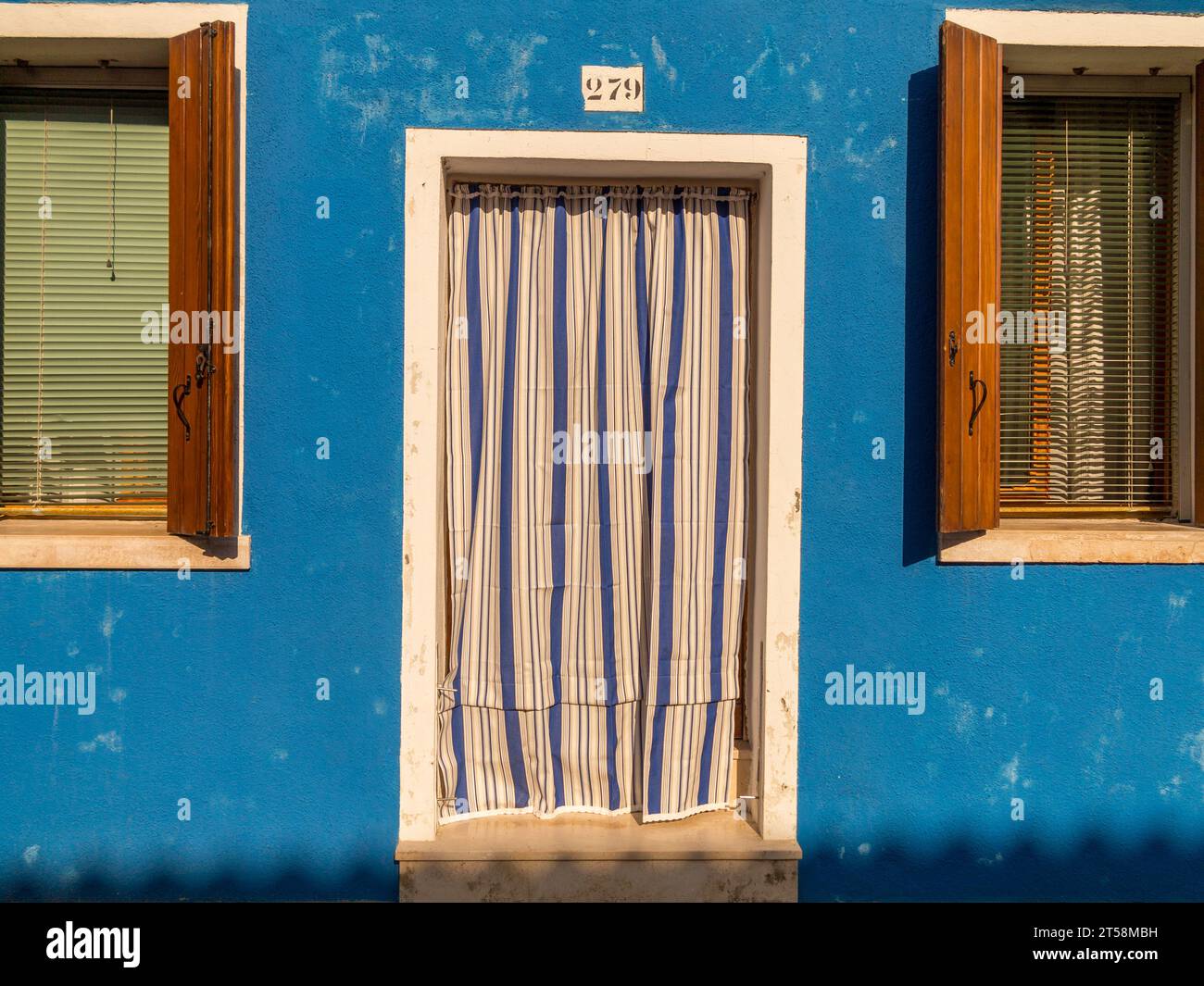 A house in Burano in Venice, Italy. A blue and white striped sheet hides the entrance. Two windows with wooden shutters and green blinds complete the Stock Photo