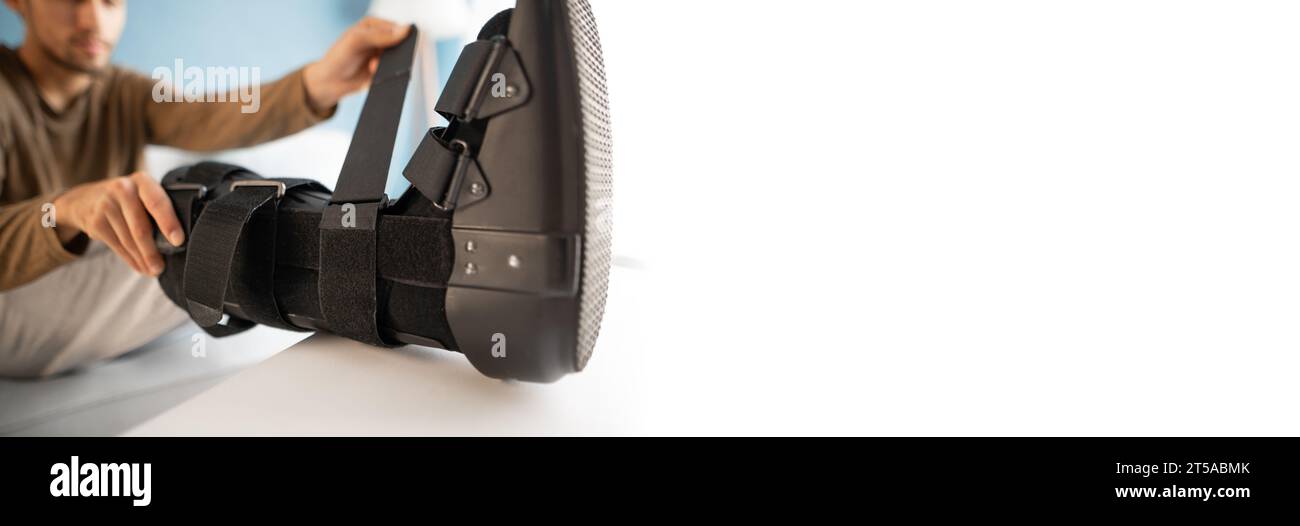 Man putting ankle sprain orthosis, black splint on leg sitting on the sofa on white background. Ankle foot orthosis. Banner. Stock Photo